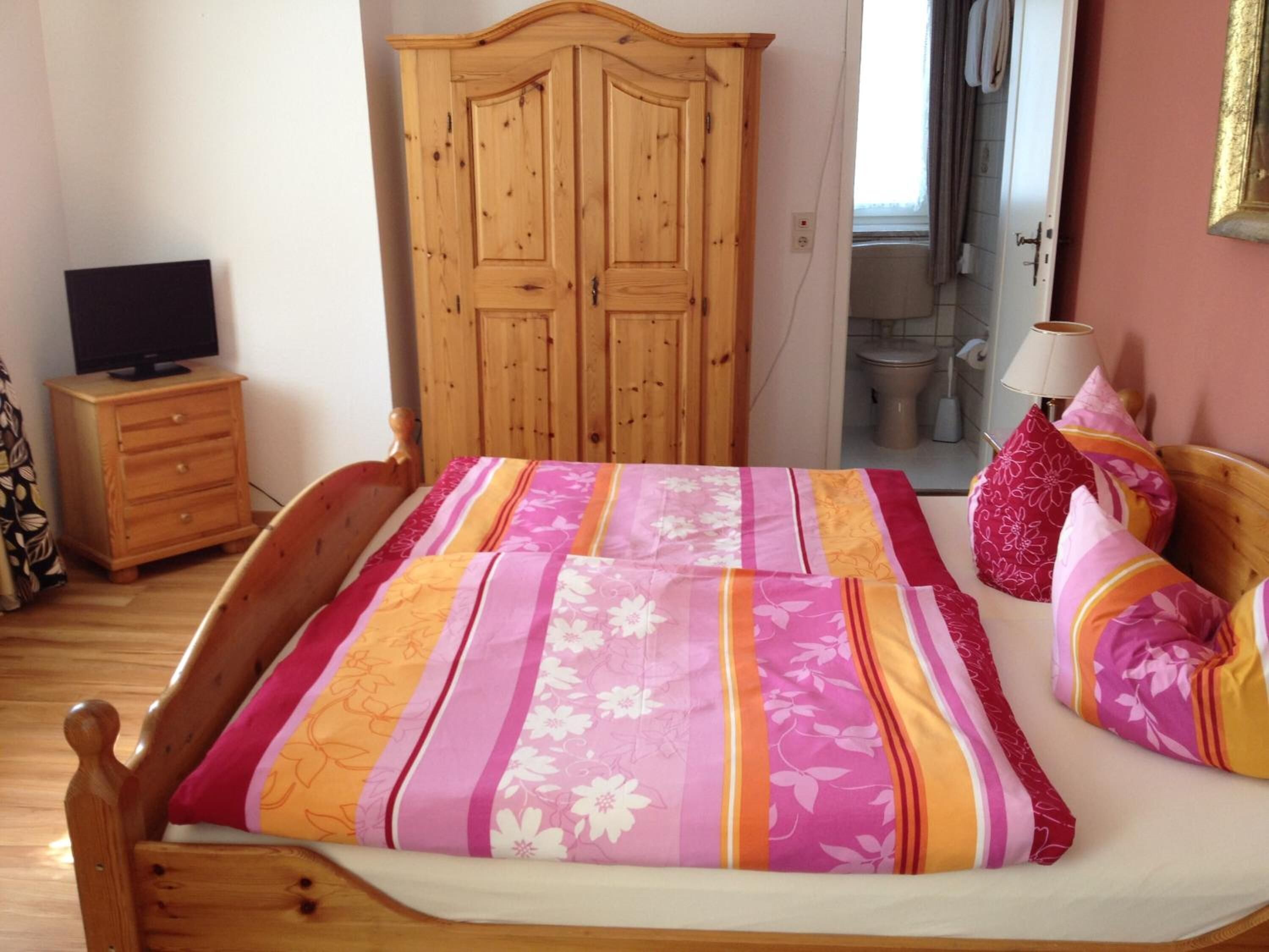 Un dormitorio con cama y armario de madera, televisor sobre un mueble de madera, y un baño visible a través de una puerta abierta.