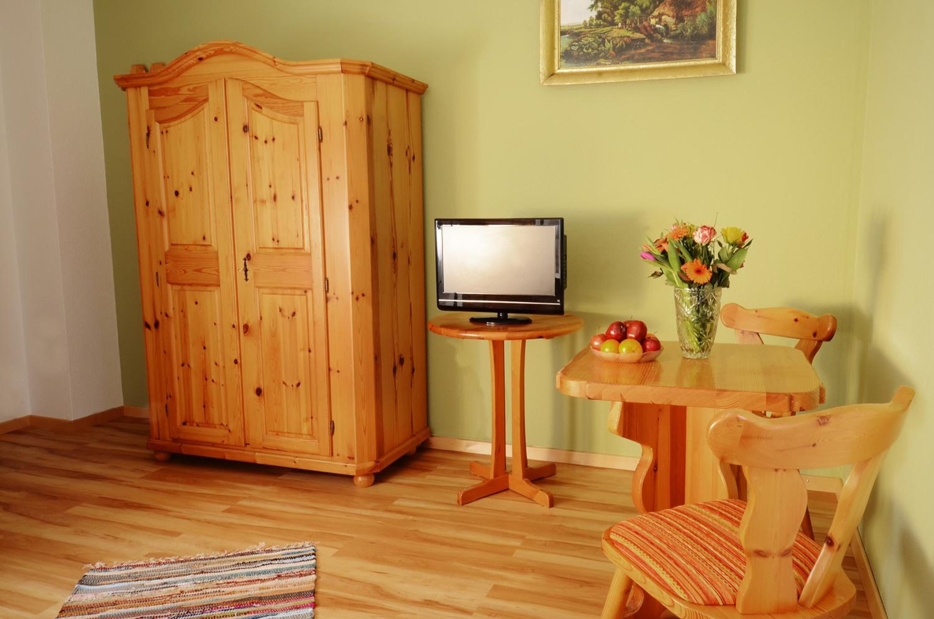 Een houten kast, een kleine tafel met een televisie, een stoel en een vaas met bloemen.