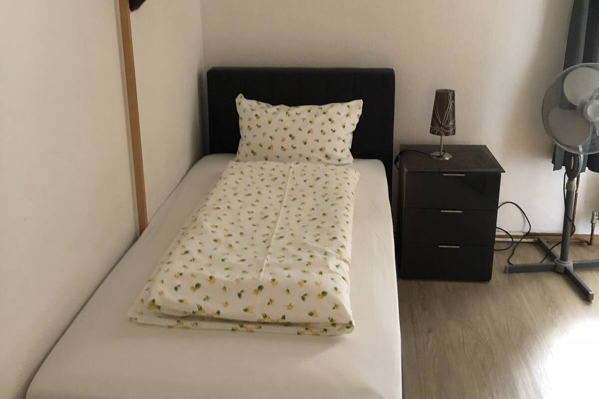 A single bed with a white mattress and a patterned blanket, a bedside table with a lamp, and a standing fan.