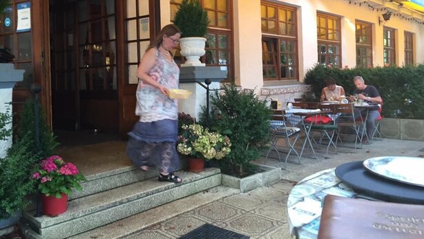 Un restaurante con terraza, una mujer caminando hacia la entrada y un hombre cenando en el interior.