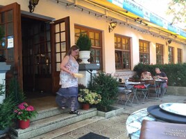Restaurante com área externa, uma mulher caminhando em direção à entrada e um homem jantando no interior.