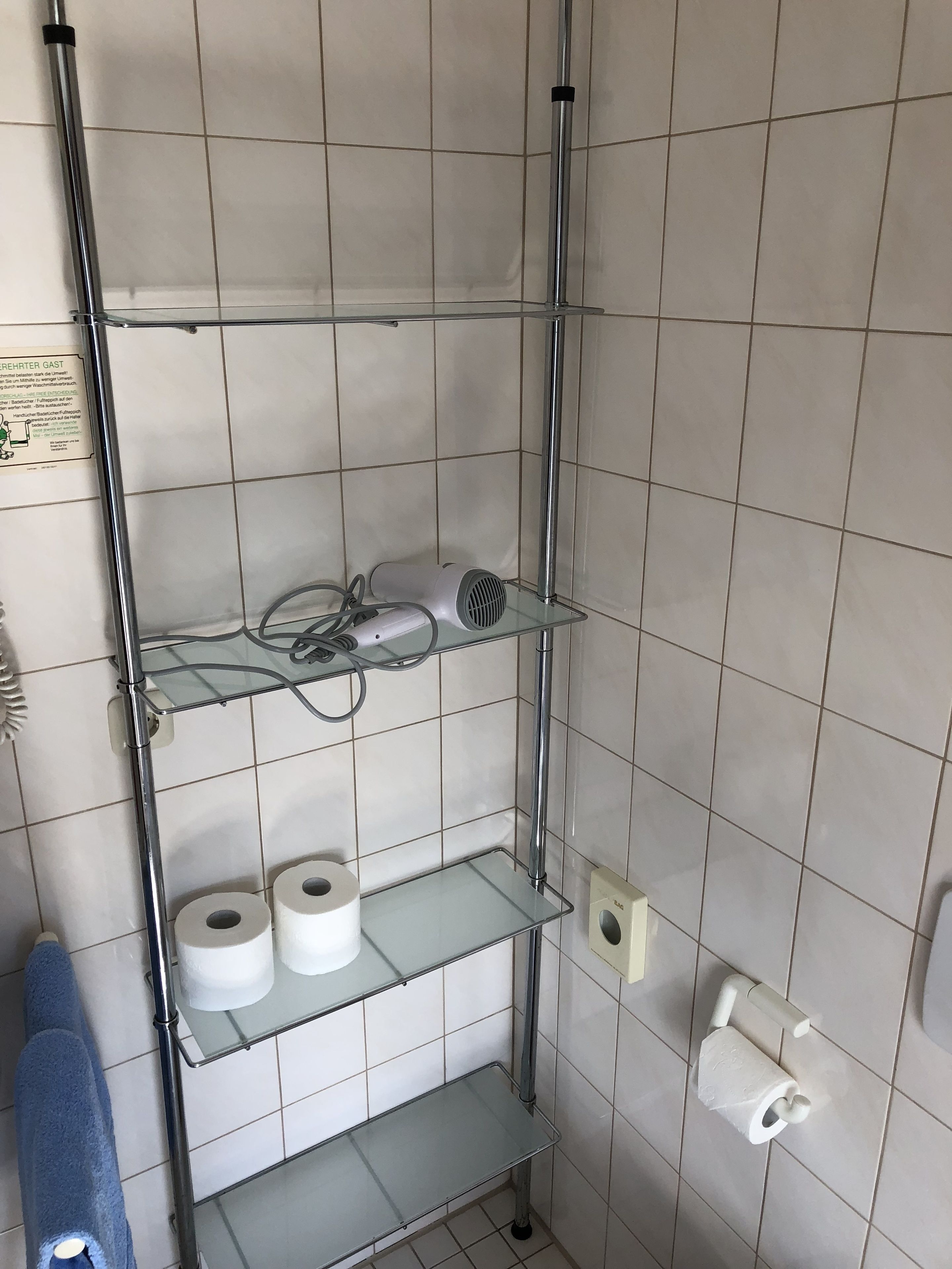 A bathroom with a tiled wall, a glass shelf holding a hairdryer, and two rolls of toilet paper on lower shelves.