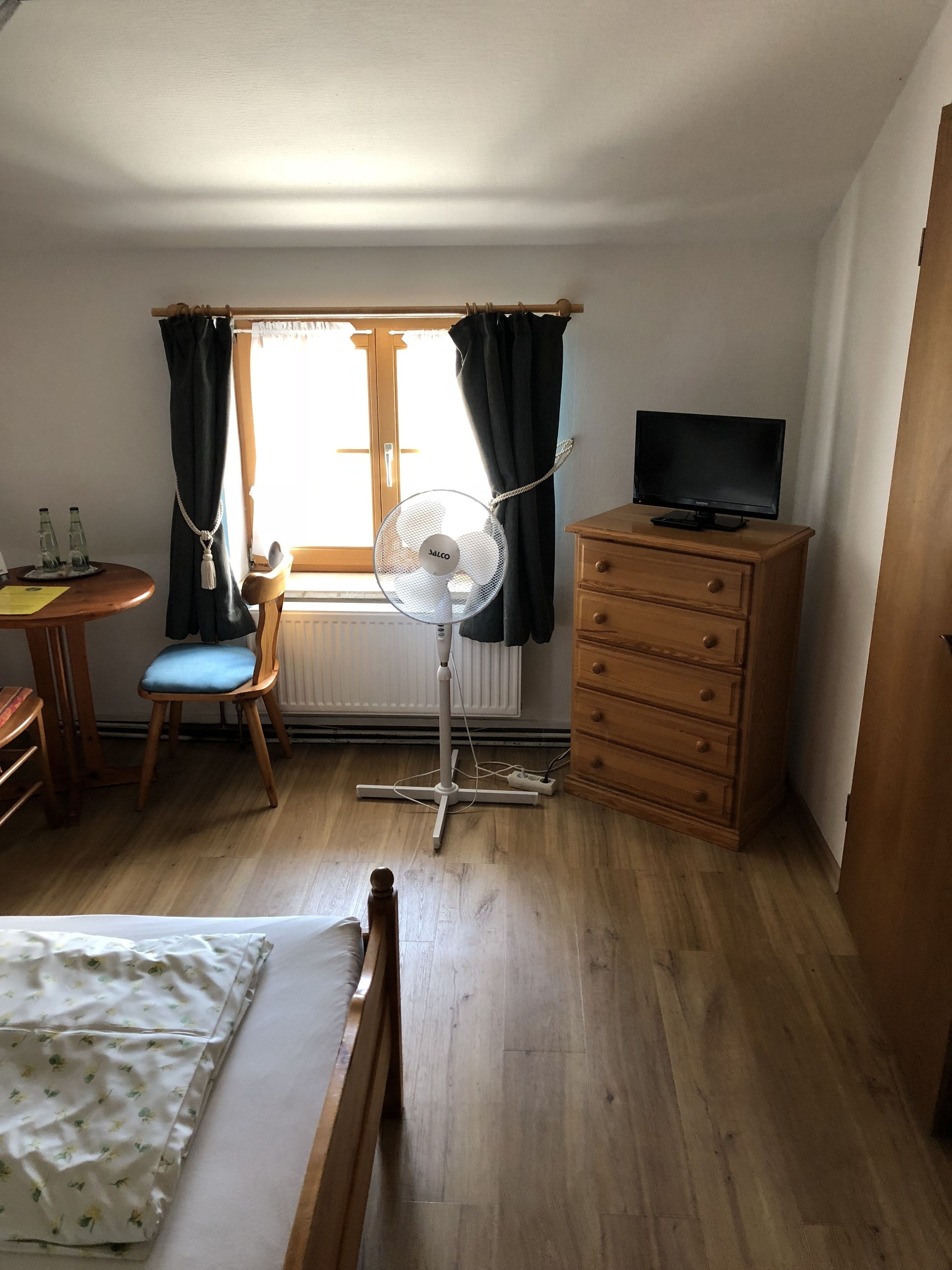 Una habitación con cama, escritorio y silla, cómoda de madera, televisor sobre un mueble bajo, ventilador de pie y ventana con cortinas.