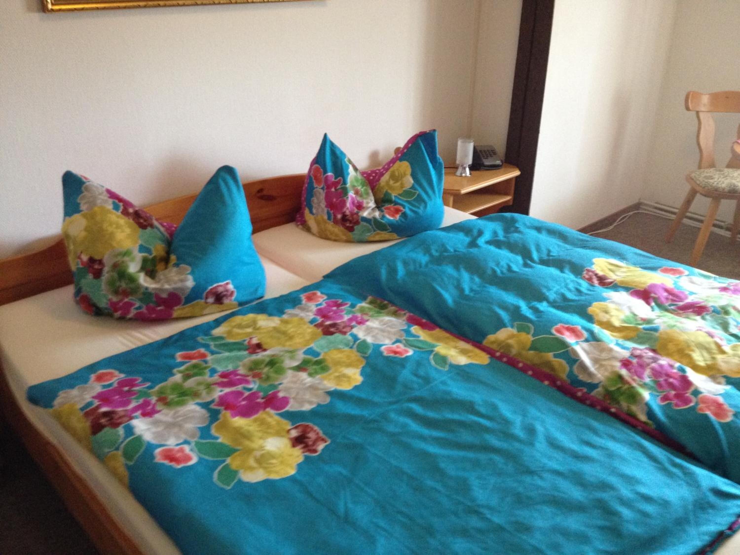 A bed with a floral bedspread and blue pillows, a wooden headboard, and a small bedside table with a lamp and a phone.