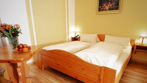 Una cama doble con ropa de cama blanca, un cabecero de madera y una mesita de noche con una lámpara. Un jarrón con flores y un frutero están sobre la mesita.