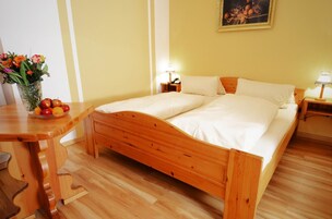 Uma cama de casal bem arrumada com lençóis brancos, um painel de madeira e uma mesa de cabeceira com uma luminária. Um vaso com flores e uma cesta de frutas estão na mesinha de cabeceira.