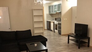 A living room with a sectional sofa, a coffee table, and a TV. There is a kitchen area with cabinets and a microwave.