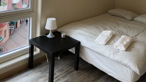 A bedroom with a large window, a bedside table with a lamp, a bed with white bedding and two towels, and a view of a building outside.