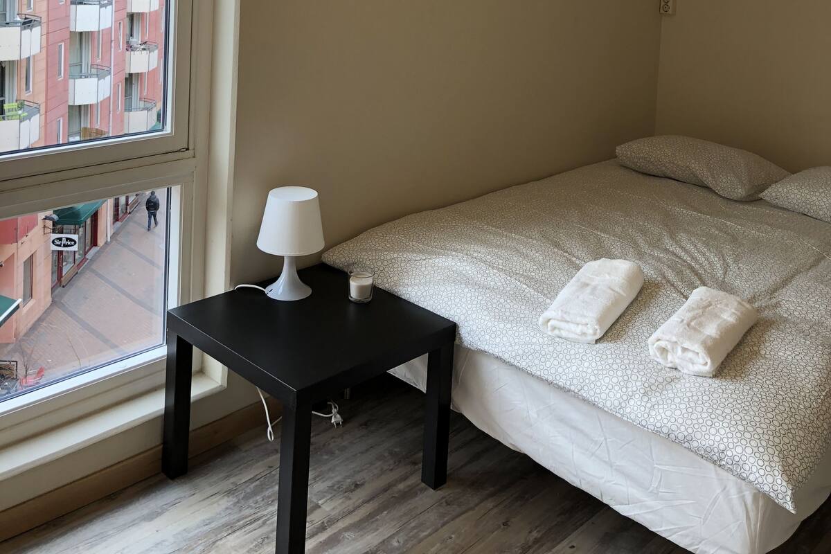A bedroom with a large window, a bedside table with a lamp, a bed with white bedding and two towels, and a view of a building outside.