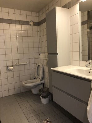 A bathroom with white tiled walls, a toilet, a sink with a mirror, and a trash bin.