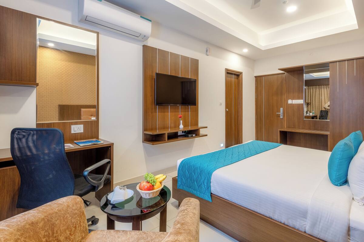 A hotel room with a bed, a desk, a chair, a TV, and a small table with fruit.