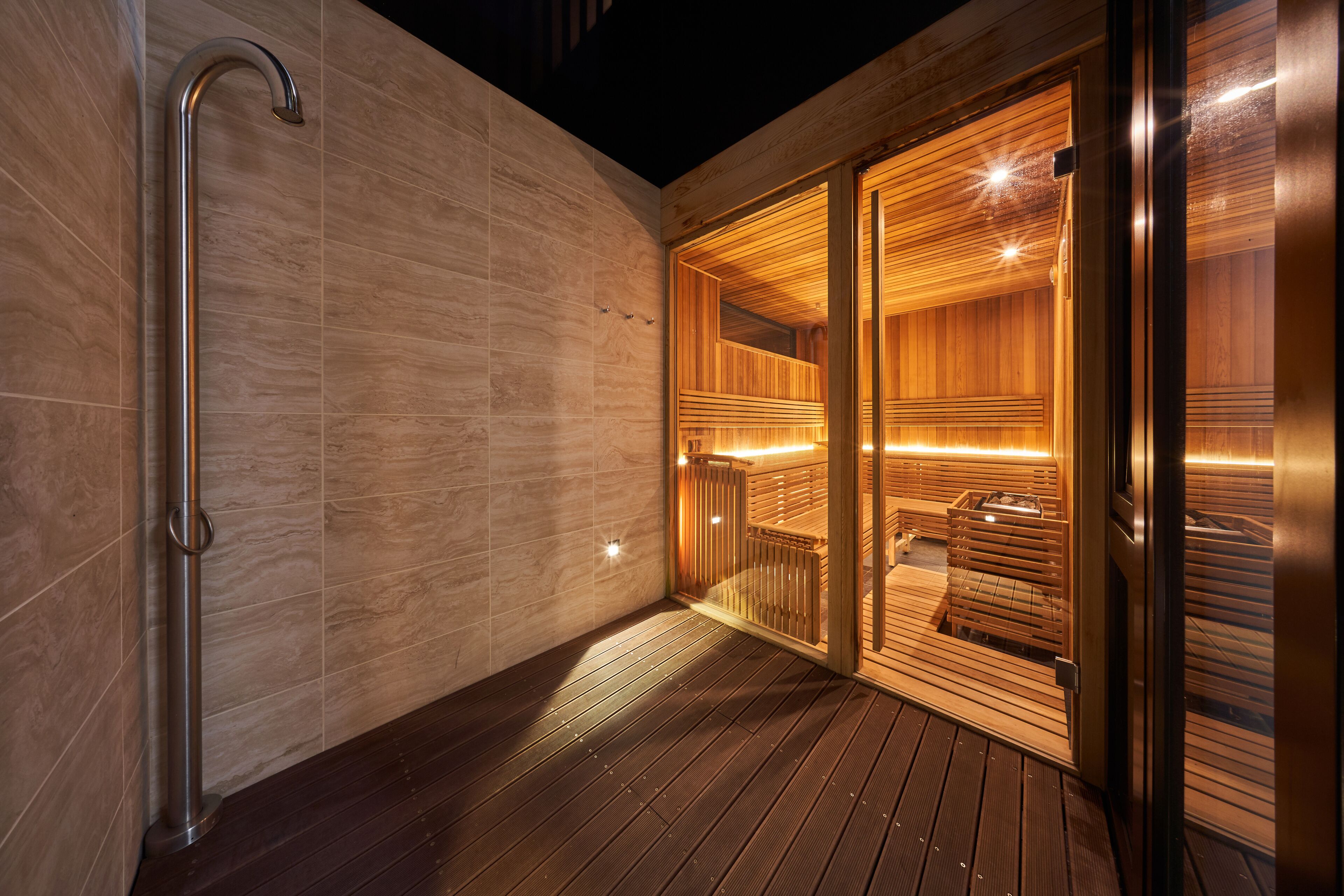 A modern sauna with wooden walls and a glass door, a metal handrail, and a wooden deck.
