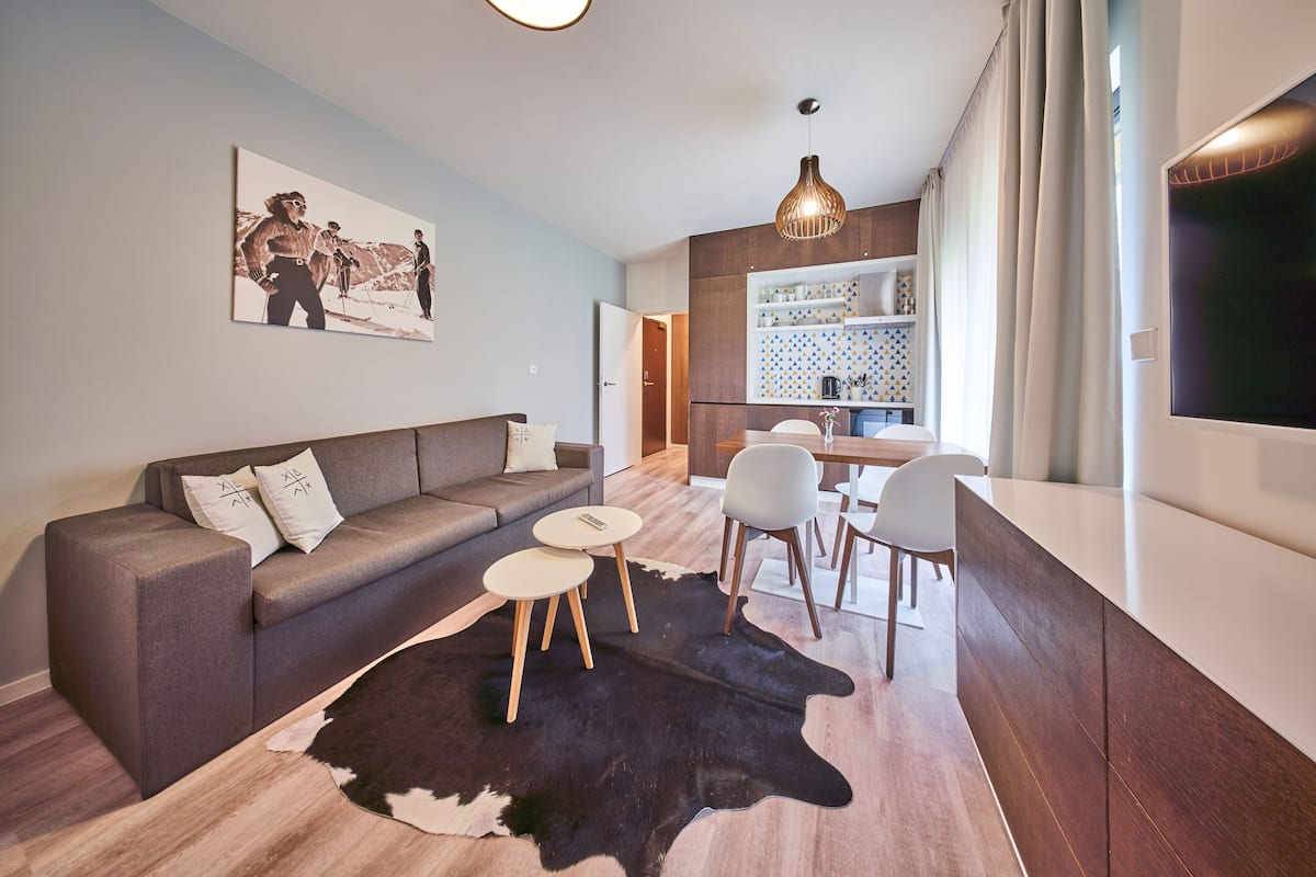 A modern living room with a sofa, a dining table with chairs, a kitchen area, and a large TV.