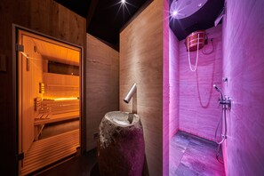 Un sauna avec un banc en bois et un bassin en pierre, un espace douche avec un seau et un tuyau d’arrosage, et une porte en bois.