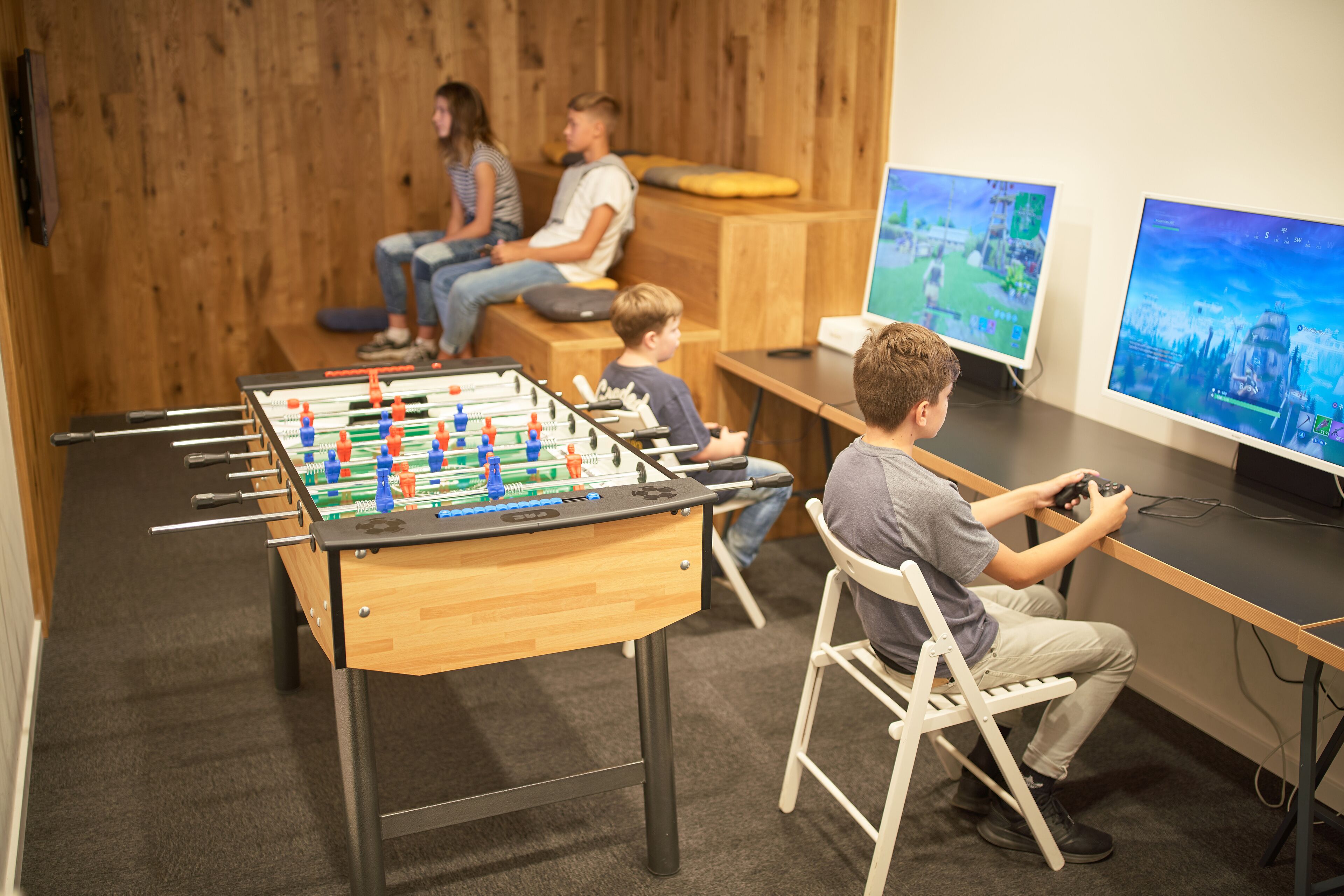 A room with a foosball table, two monitors displaying video games, and a seating area with people.