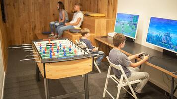 Habitación con mesa de fútbol de mesa, dos monitores mostrando videojuegos y una zona de estar con gente.