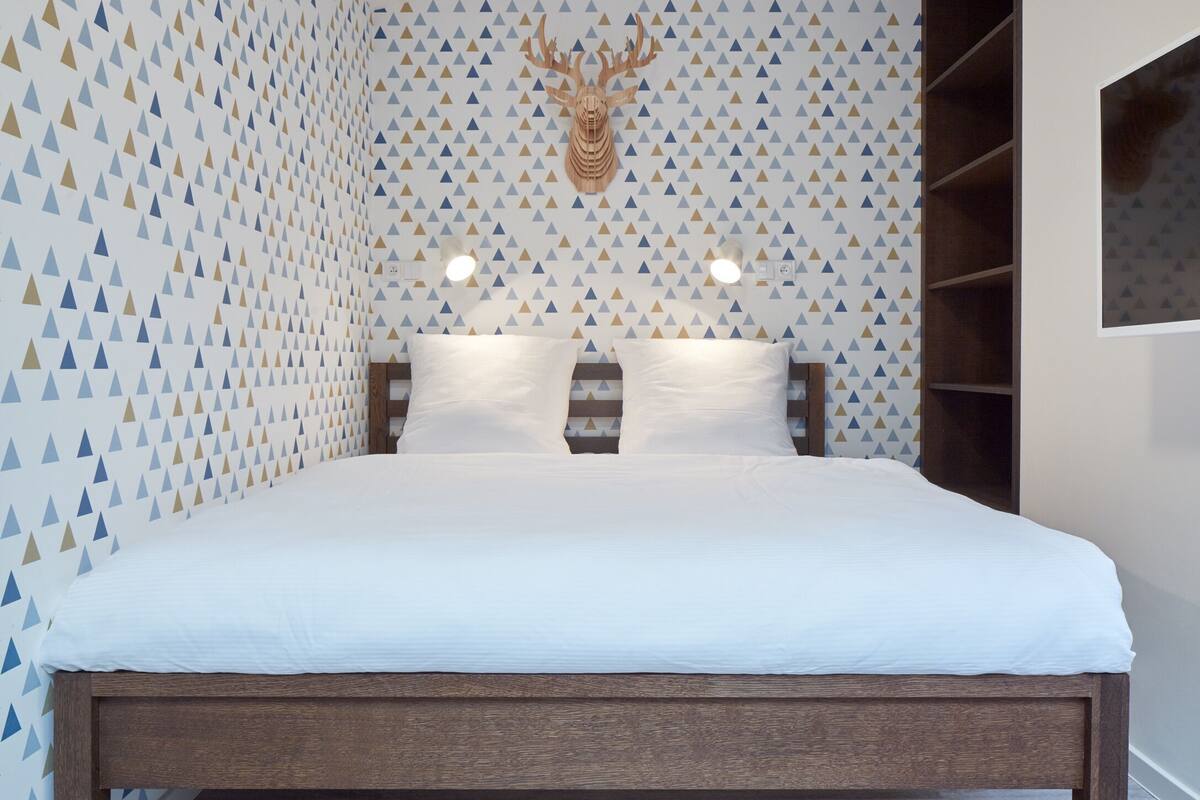 A bedroom with a patterned wall, a mounted deer head, a bed with white bedding, and a built-in shelf unit.