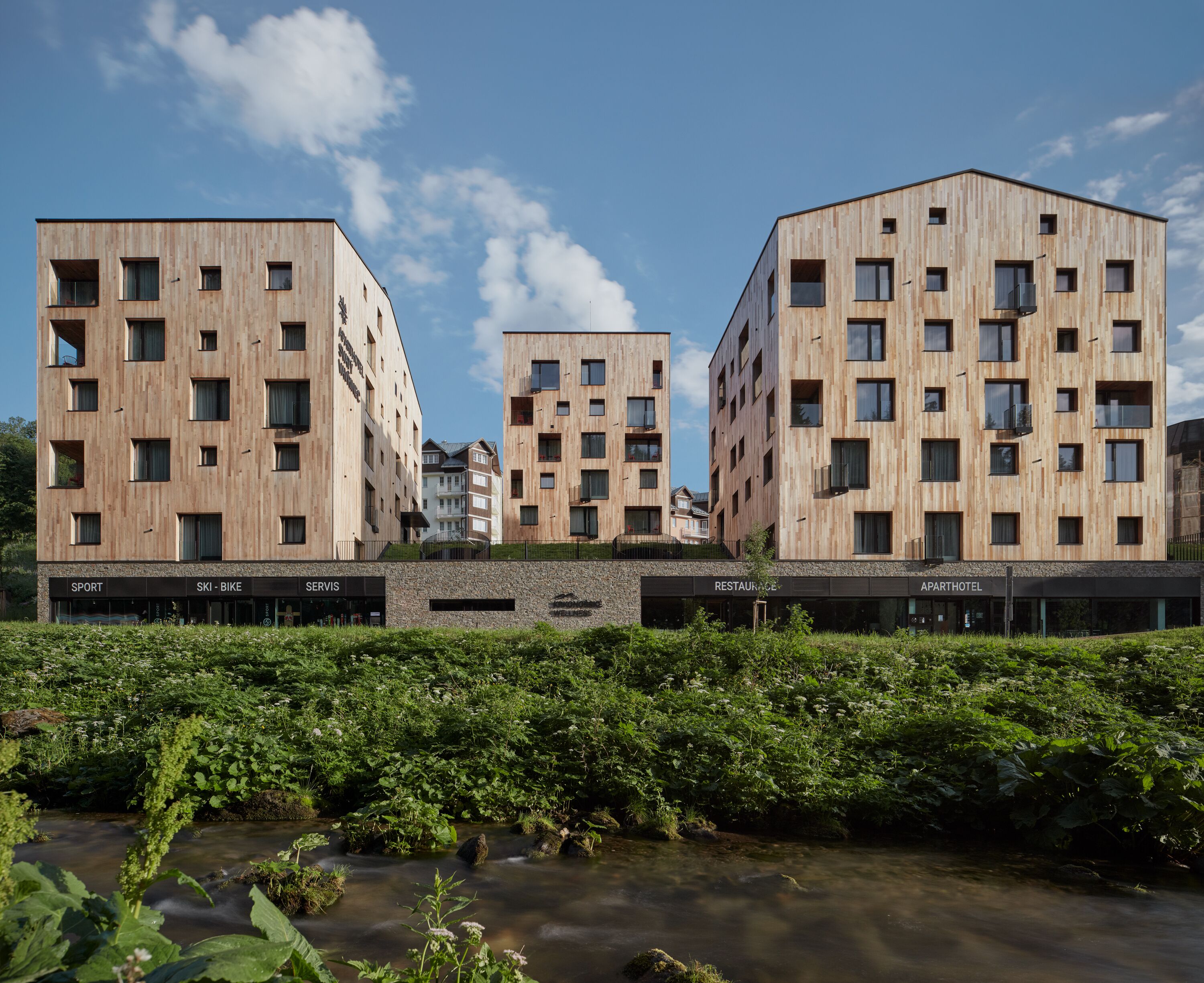 A modern building with multiple floors, featuring a mix of rectangular and square windows. In front of the building, there is a landscaped area with a small stream and lush greenery.