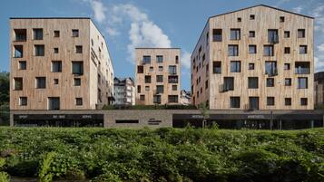 A modern building with multiple floors, featuring a mix of rectangular and square windows. In front of the building, there is a landscaped area with a small stream and lush greenery.