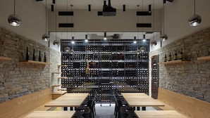 Un comedor moderno con una larga mesa de madera y sillas negras, una pared de piedra con botellas de vino e iluminación colgante.