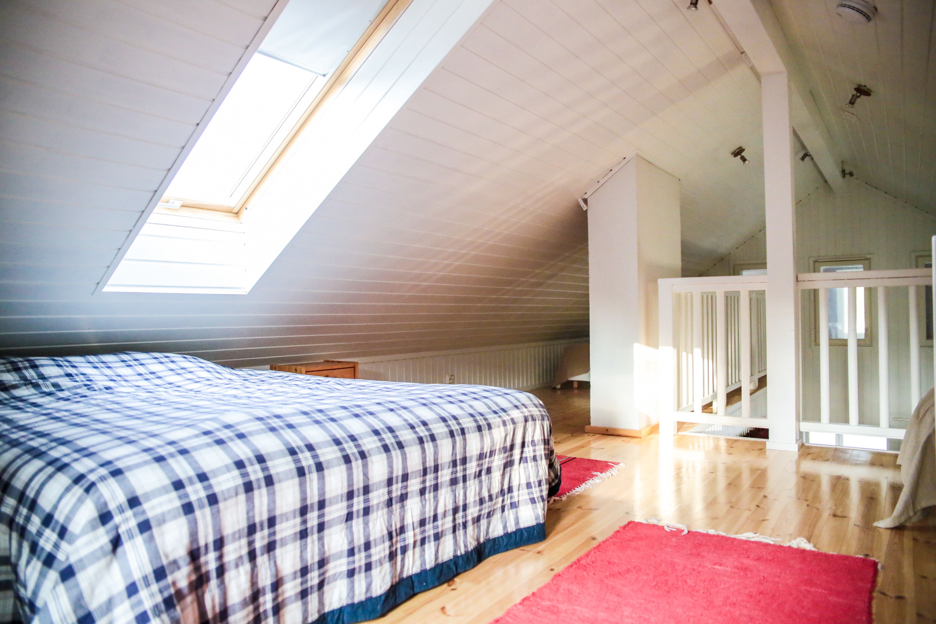 Ein Schlafzimmer mit schrägen Wänden, einem Oberlicht, einem Bett mit karierter Tagesdecke, einem roten Teppich und einem weißen Geländer.