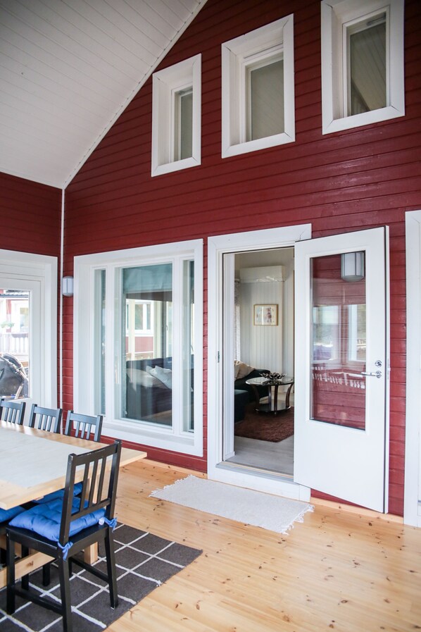 A red exterior with white-framed windows and a sliding glass door leading to an interior space with a dining table, chairs, and a sofa. - Rantakari Cottage (Kotka)