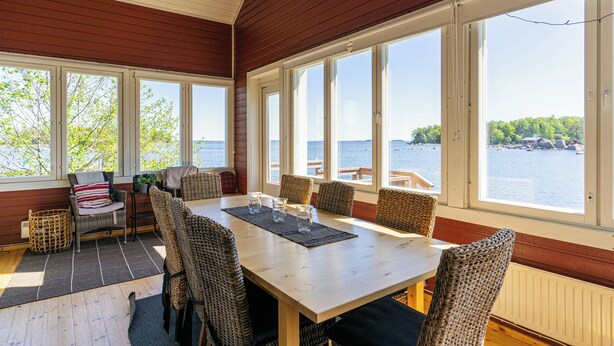 A dining area with a wooden table and wicker chairs, large windows with a view of the water and trees.