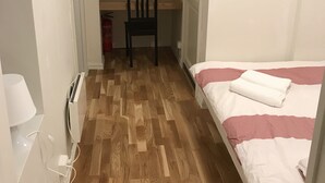 A bedroom with a wooden parquet floor, a bed with pink and white bedding, a chair, and a desk with a red object on it.