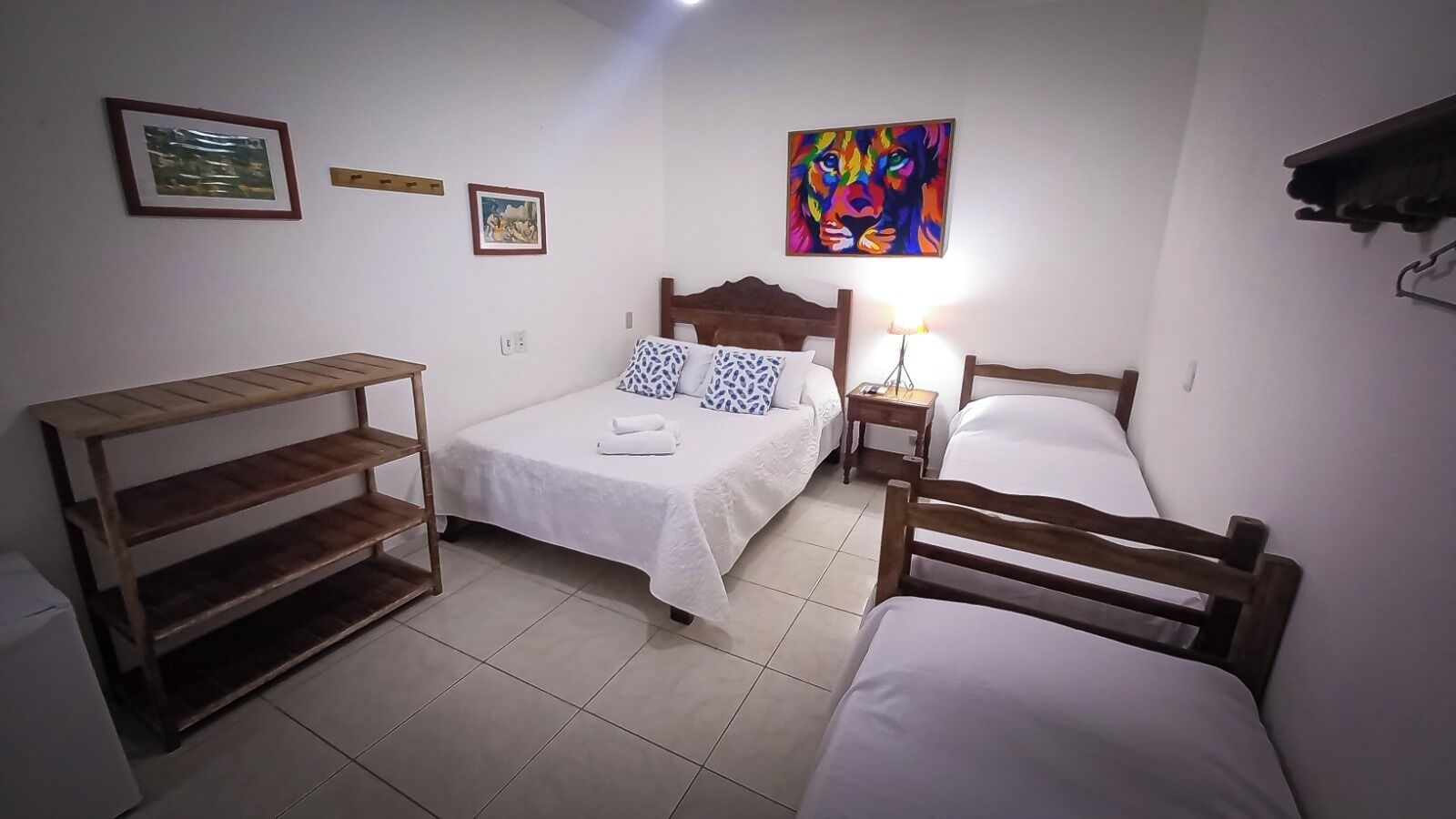 A bedroom with two single beds, a wooden shelf unit, a nightstand with a lamp, and a colorful painting on the wall.