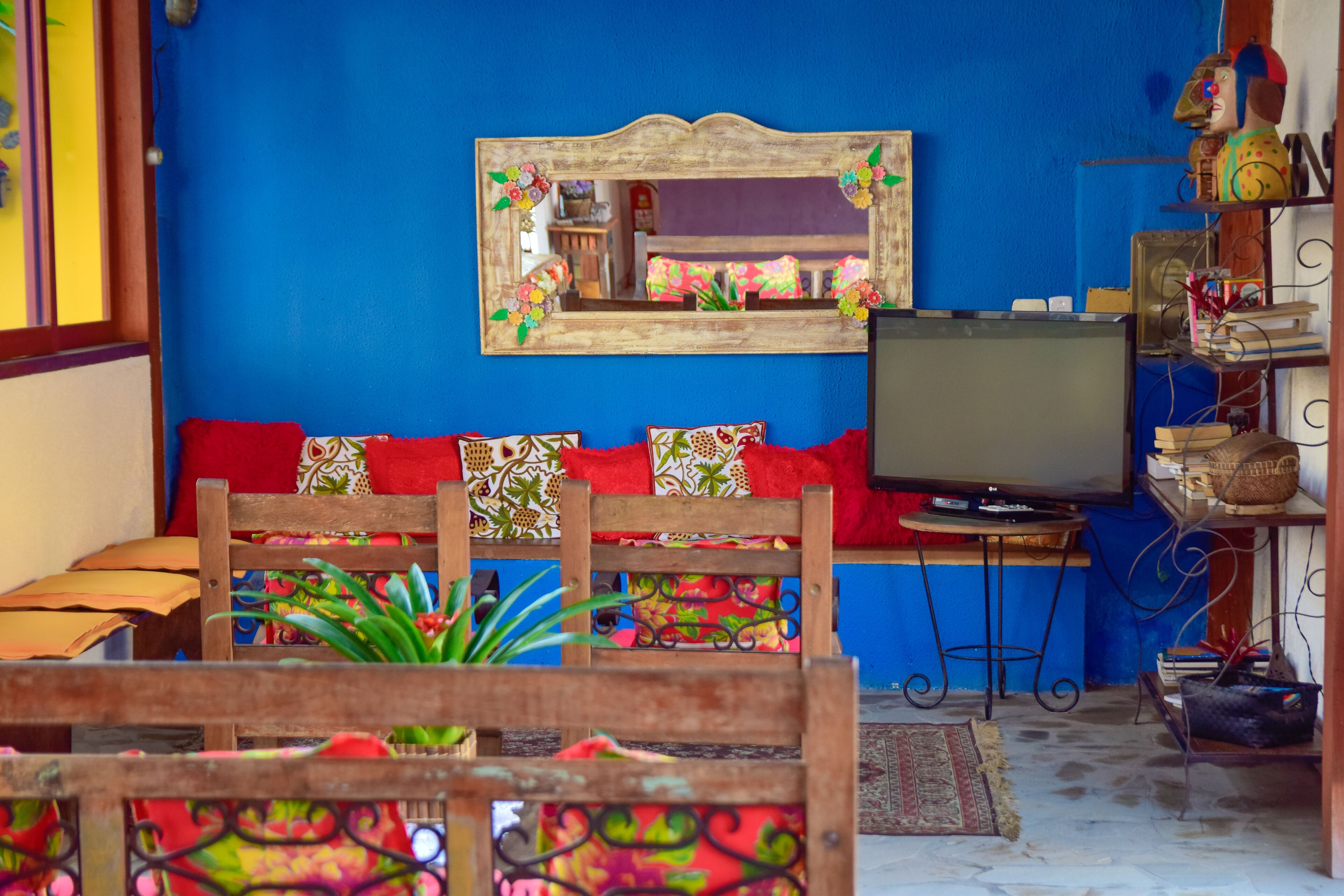 A room with a blue wall, a wooden bench with colorful cushions, a television on a stand, and a shelf with various items.