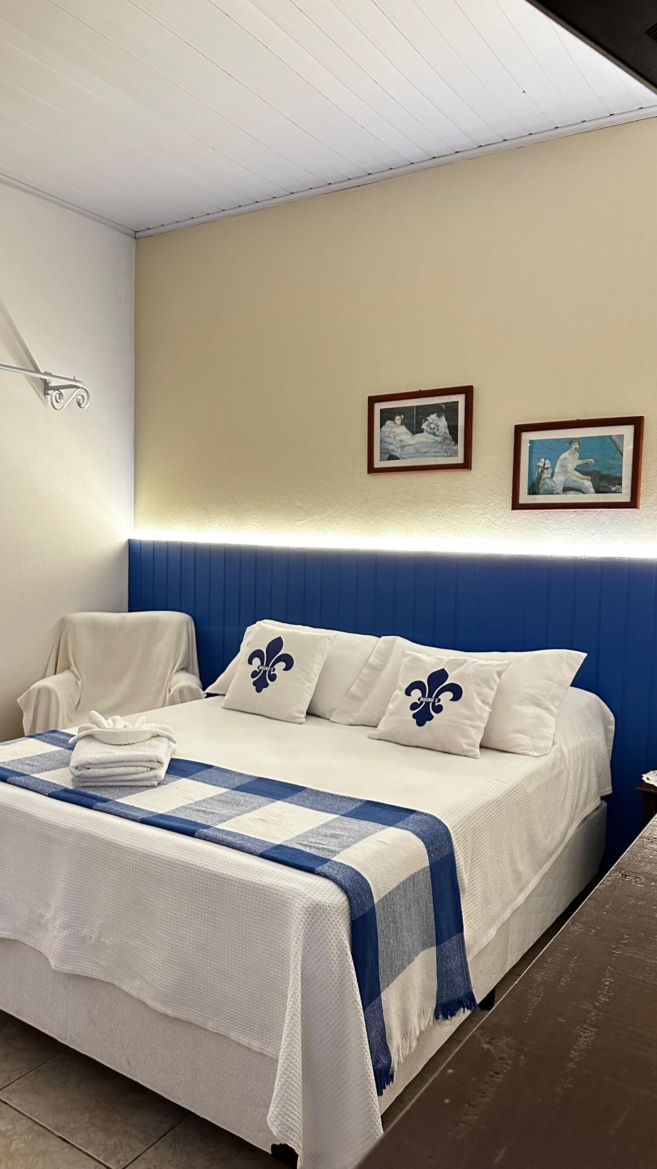 A neatly made bed with a checkered blanket, a pillow with a fleur-de-lis design, and two framed pictures on the wall.
