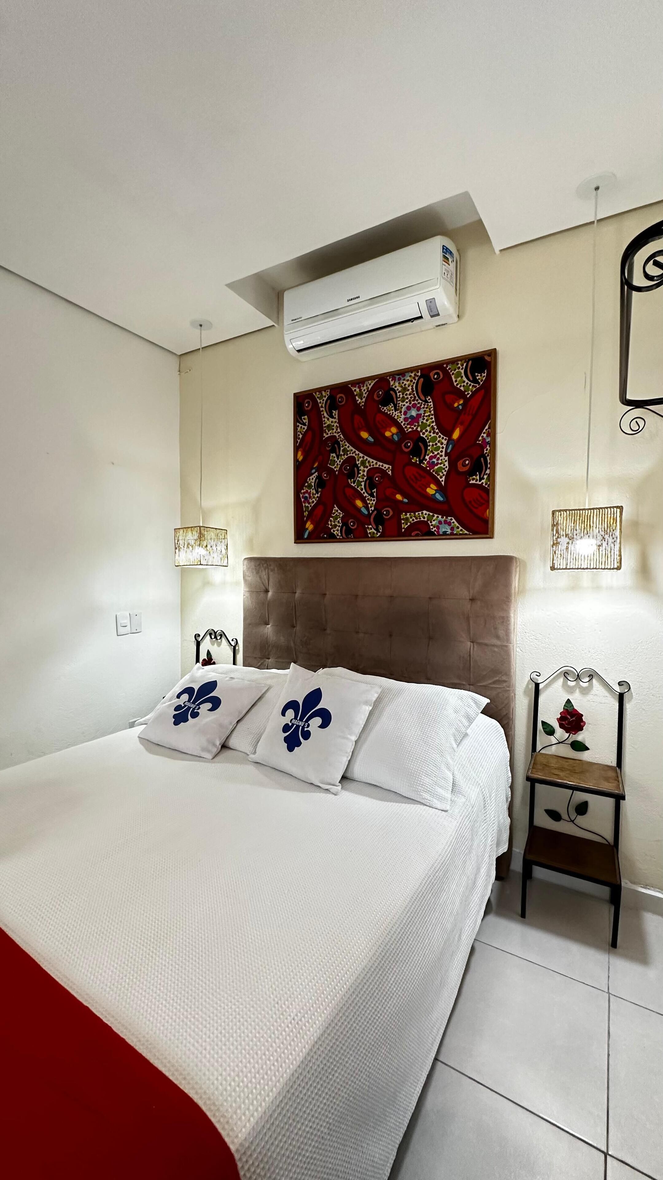 A bedroom with a bed, an air conditioner, a decorative painting, a bedside lamp, and a small wooden chair.
