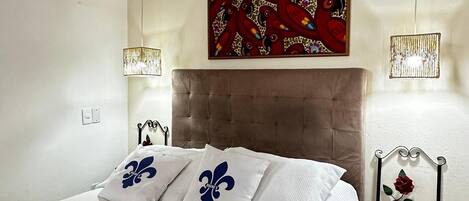 Un dormitorio con cama, aire acondicionado, un cuadro decorativo, una lámpara de noche y una pequeña silla de madera.