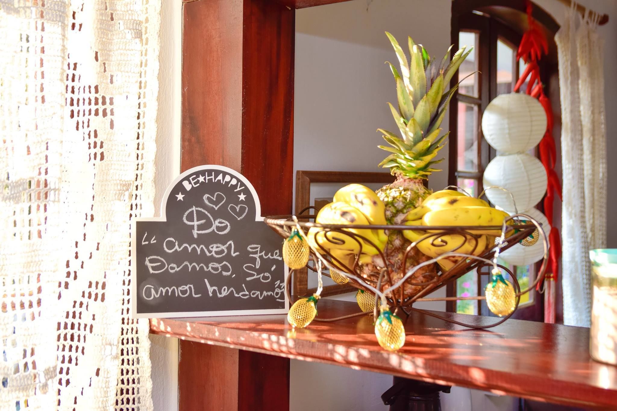 A wooden table with a fruit basket containing bananas and a pineapple, a chalkboard sign with a message, and decorative paper lanterns.