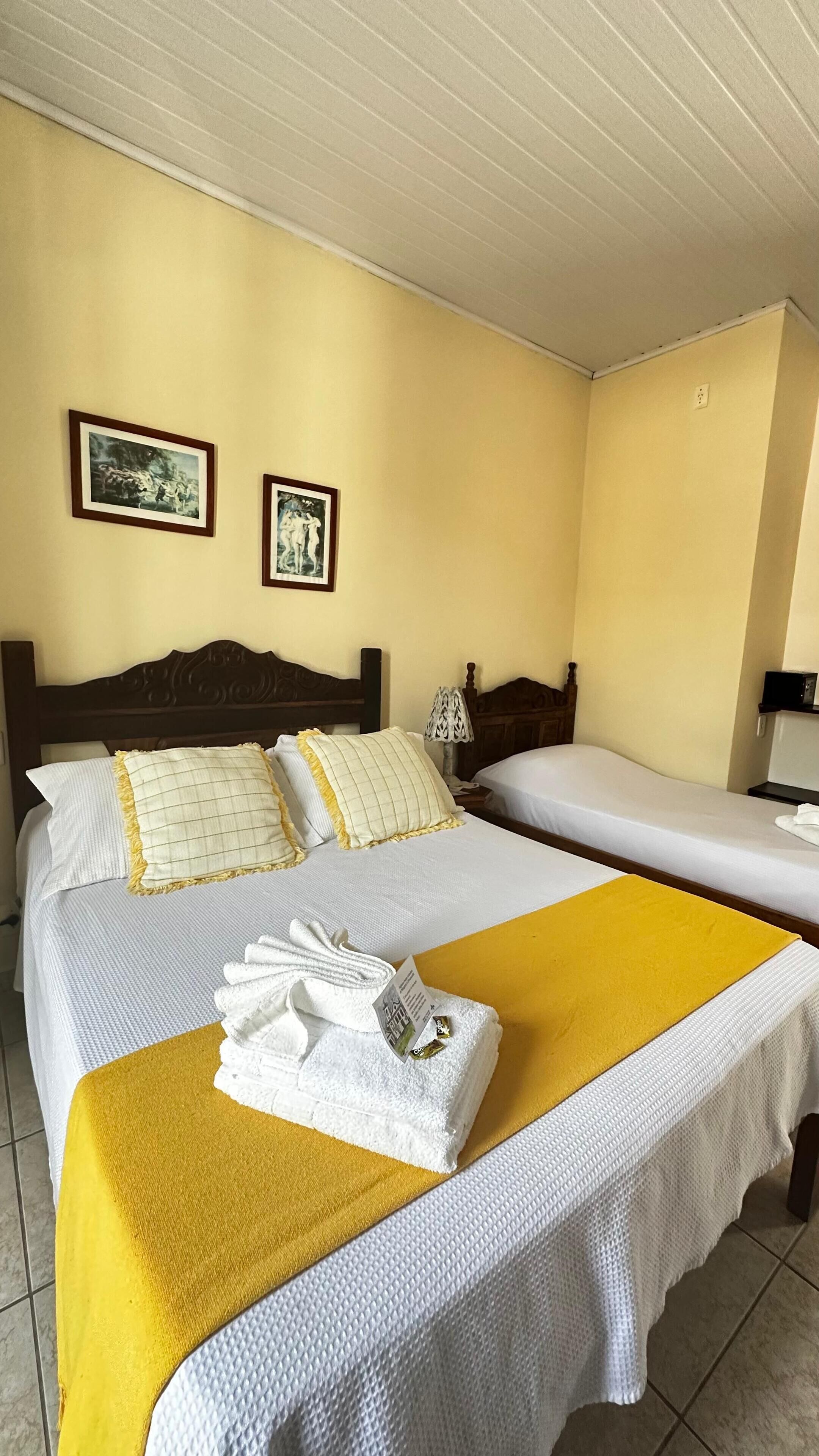A hotel room with two beds, a wooden headboard, a yellow throw blanket, and two framed pictures on the wall.