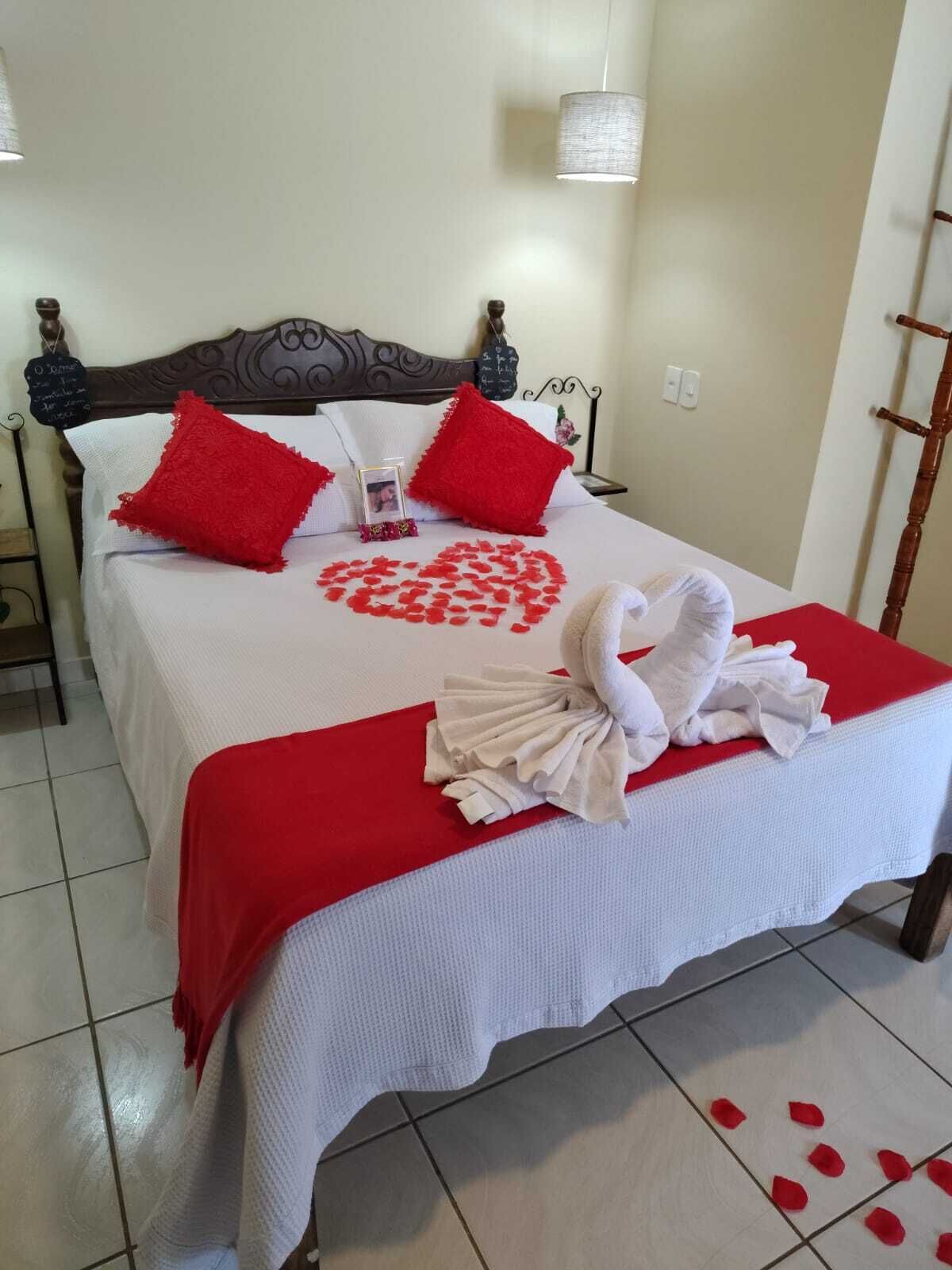 A neatly made bed with a heart-shaped arrangement of rose petals, red pillows, and a towel sculpture.
