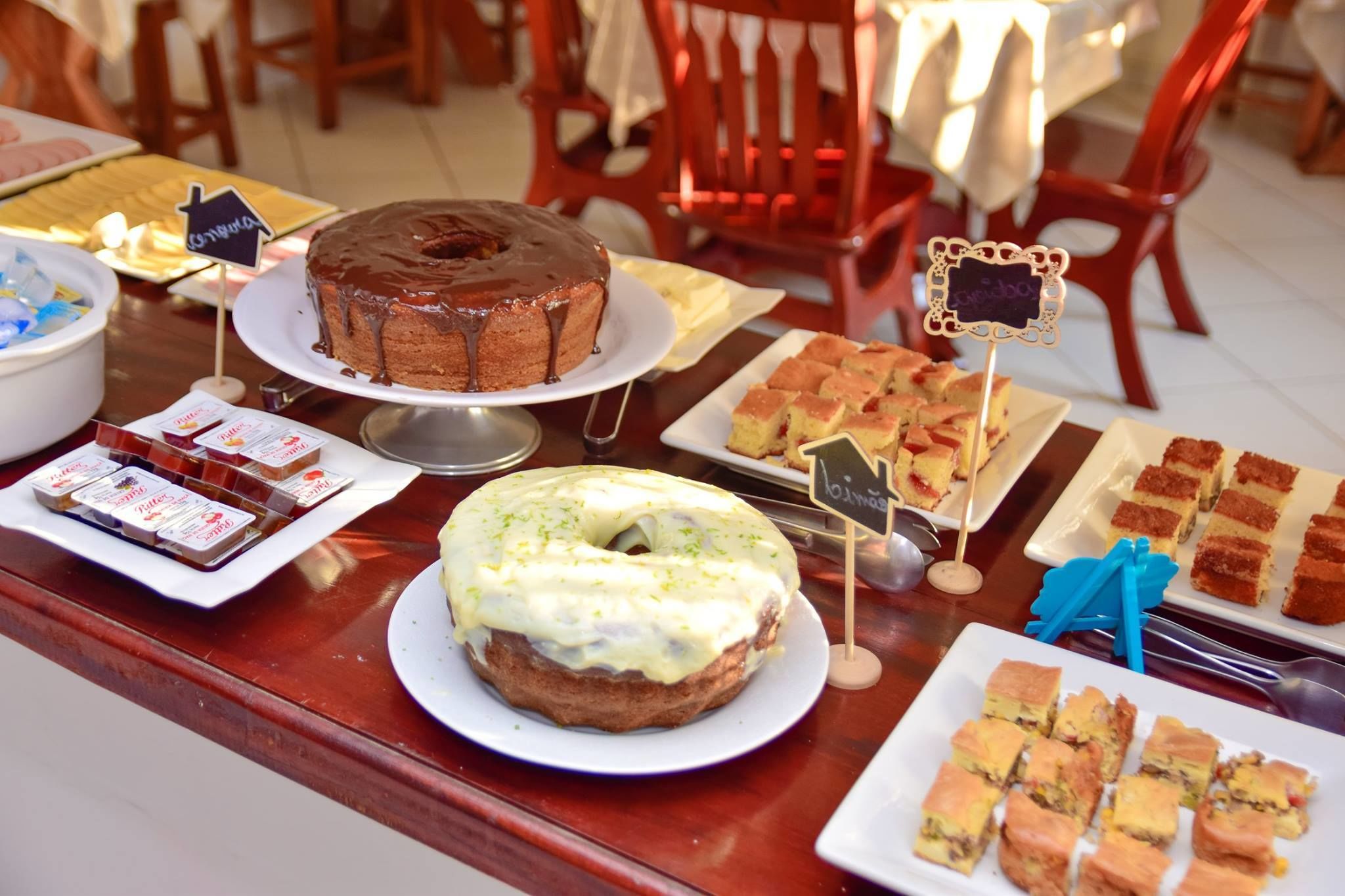 Una mesa de bufé con diversos postres, incluyendo un pastel de chocolate, un donut glaseado y varias pastas cuadradas.