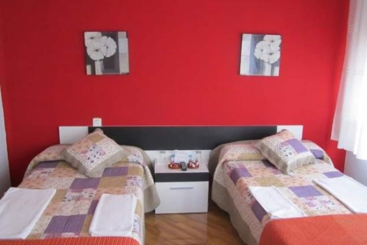 A room with two beds, each with a quilted bedspread and pillows, against a red wall with two framed pictures.