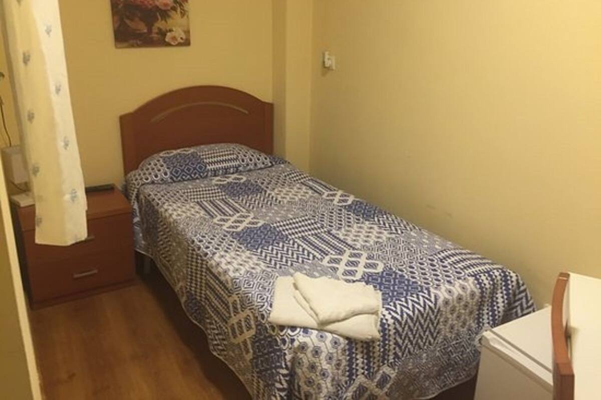 A small, single-bed room with a patterned bedspread, a wooden headboard, a nightstand, and a white appliance.