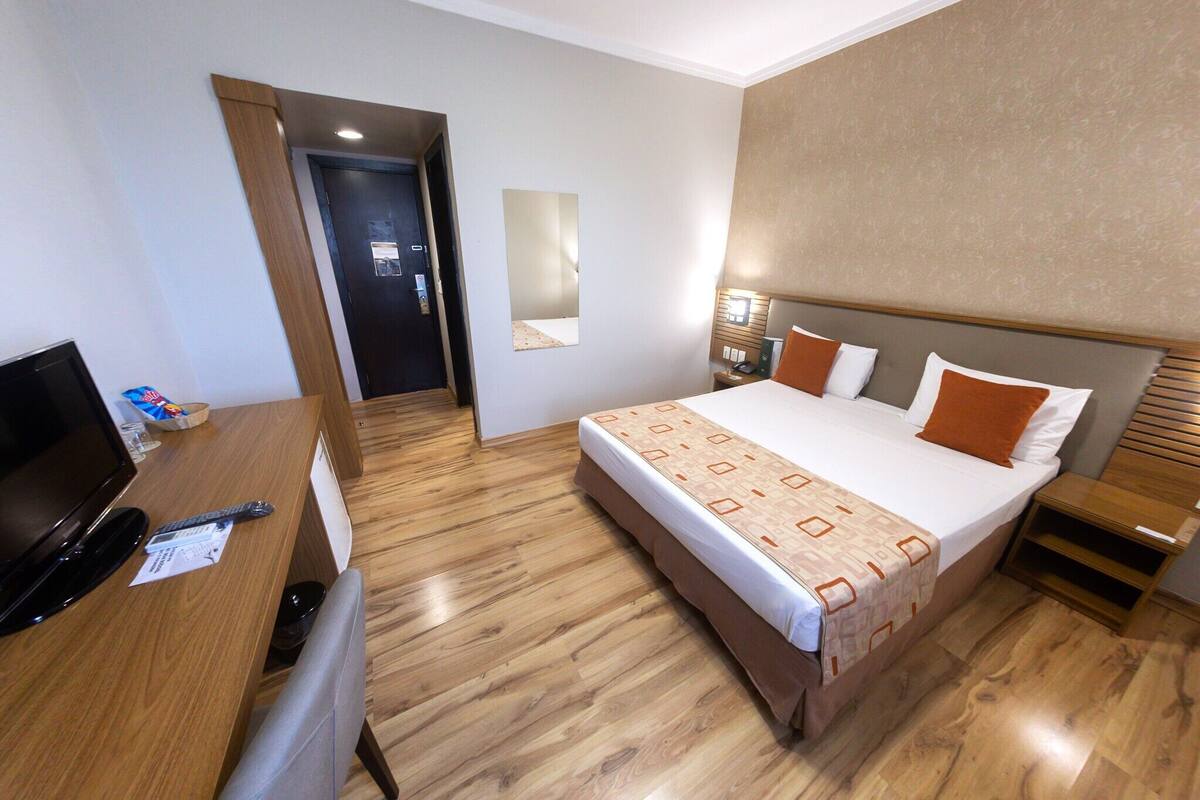 Une chambre d’hôtel comprenant un lit, une table de chevet, une lampe, un bureau avec un ordinateur et une télévision.