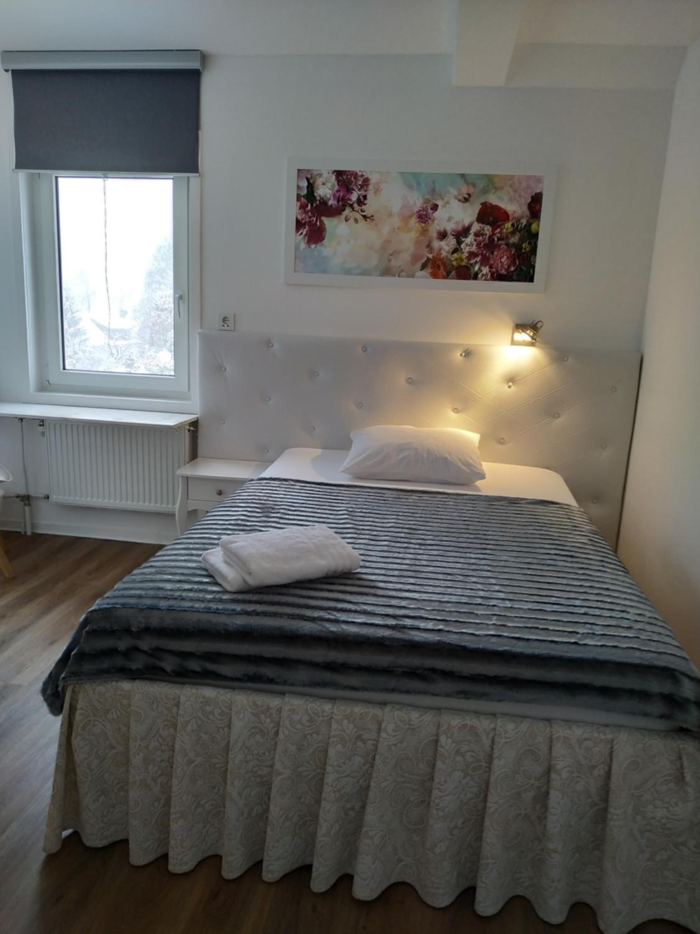 Makuuhuone, jossa on suuri sänky, ikkunasta avautuva näkymä, patteri, yöpöytä ja seinään kiinnitetty valaisin.