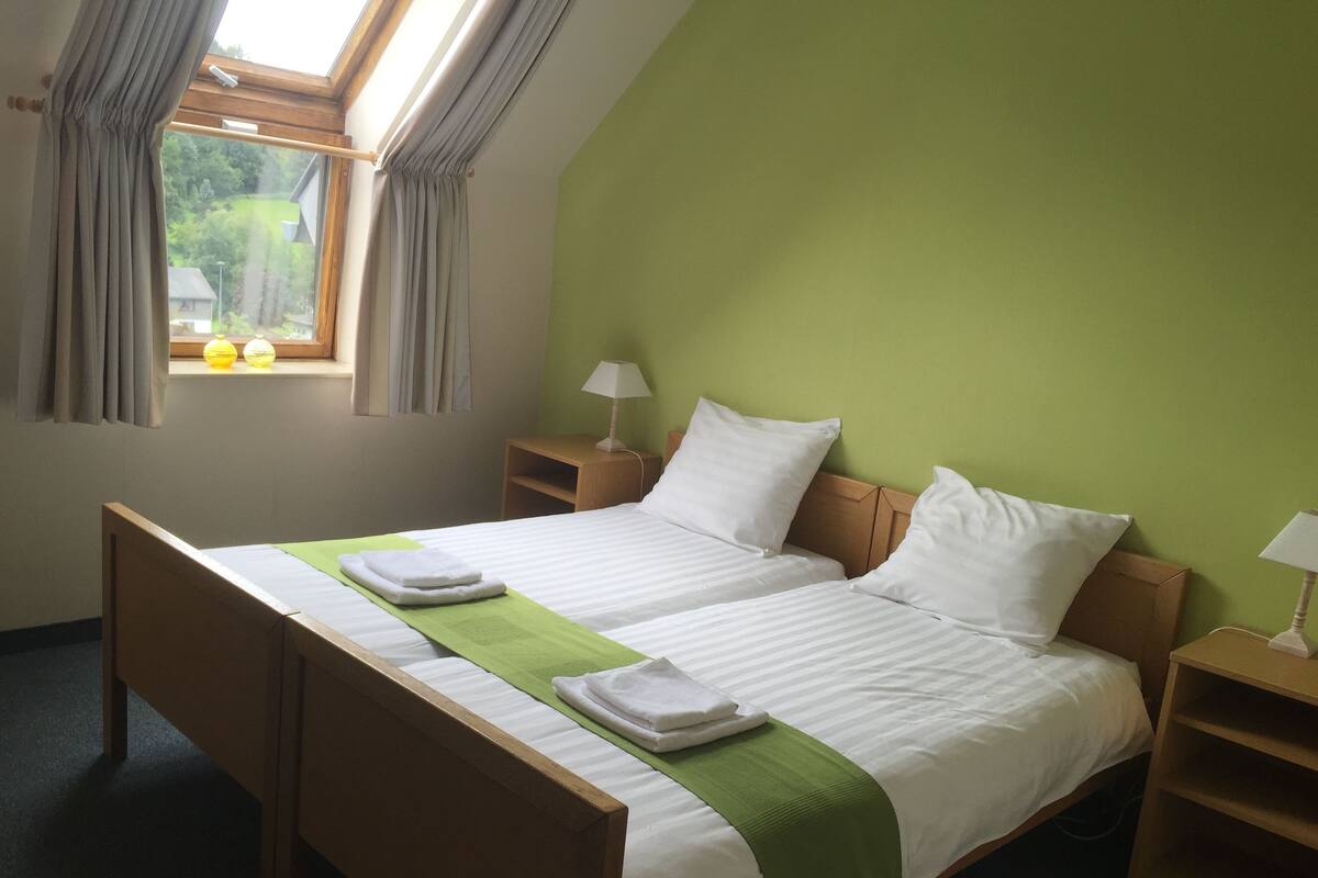 A double bed with a green bedspread, two bedside tables with lamps, a window with curtains, and a view of greenery outside.