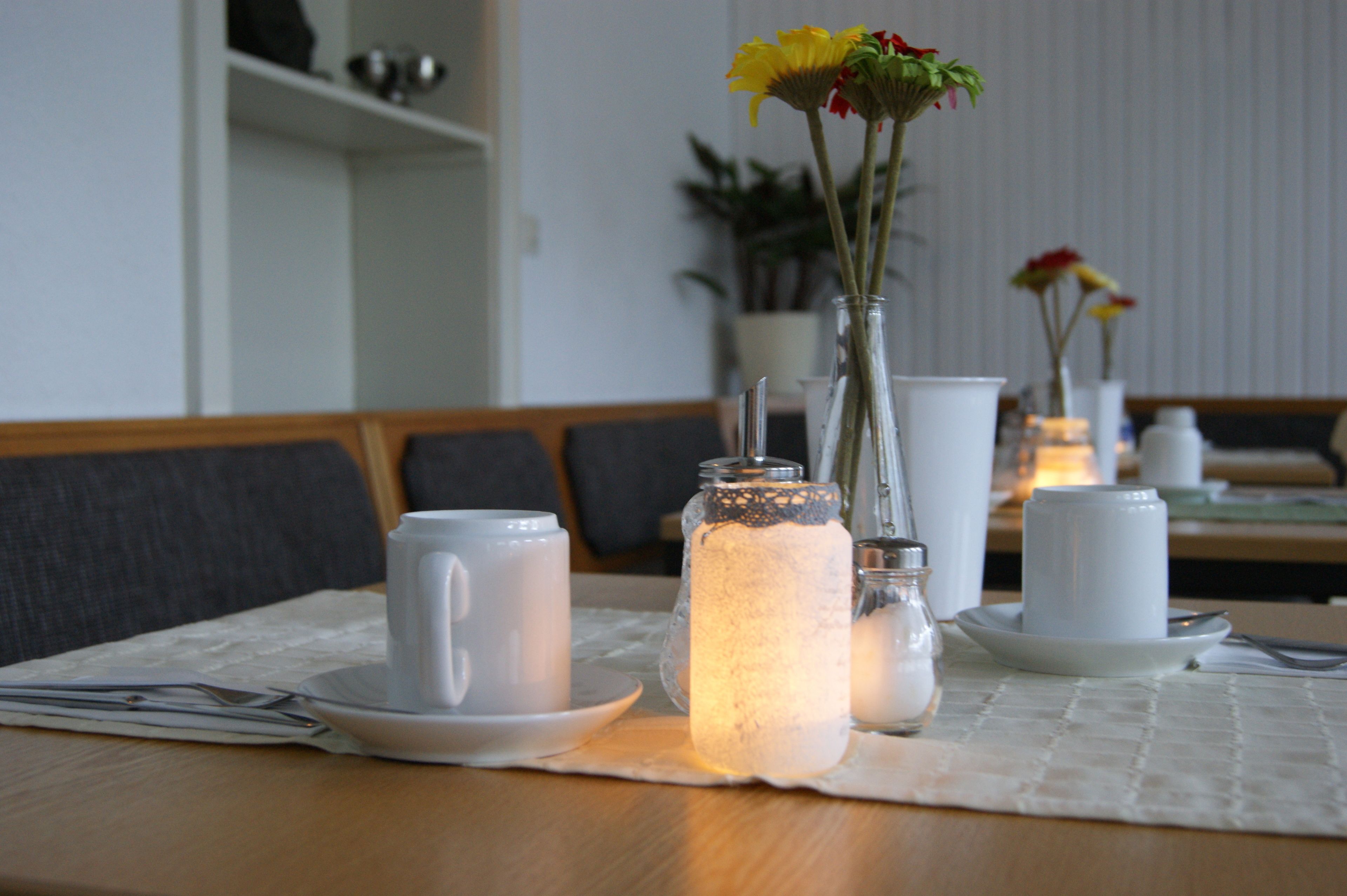 Une table à manger avec un set de table blanc, un bocal en verre contenant une bougie, une tasse blanche et un petit vase avec des fleurs.