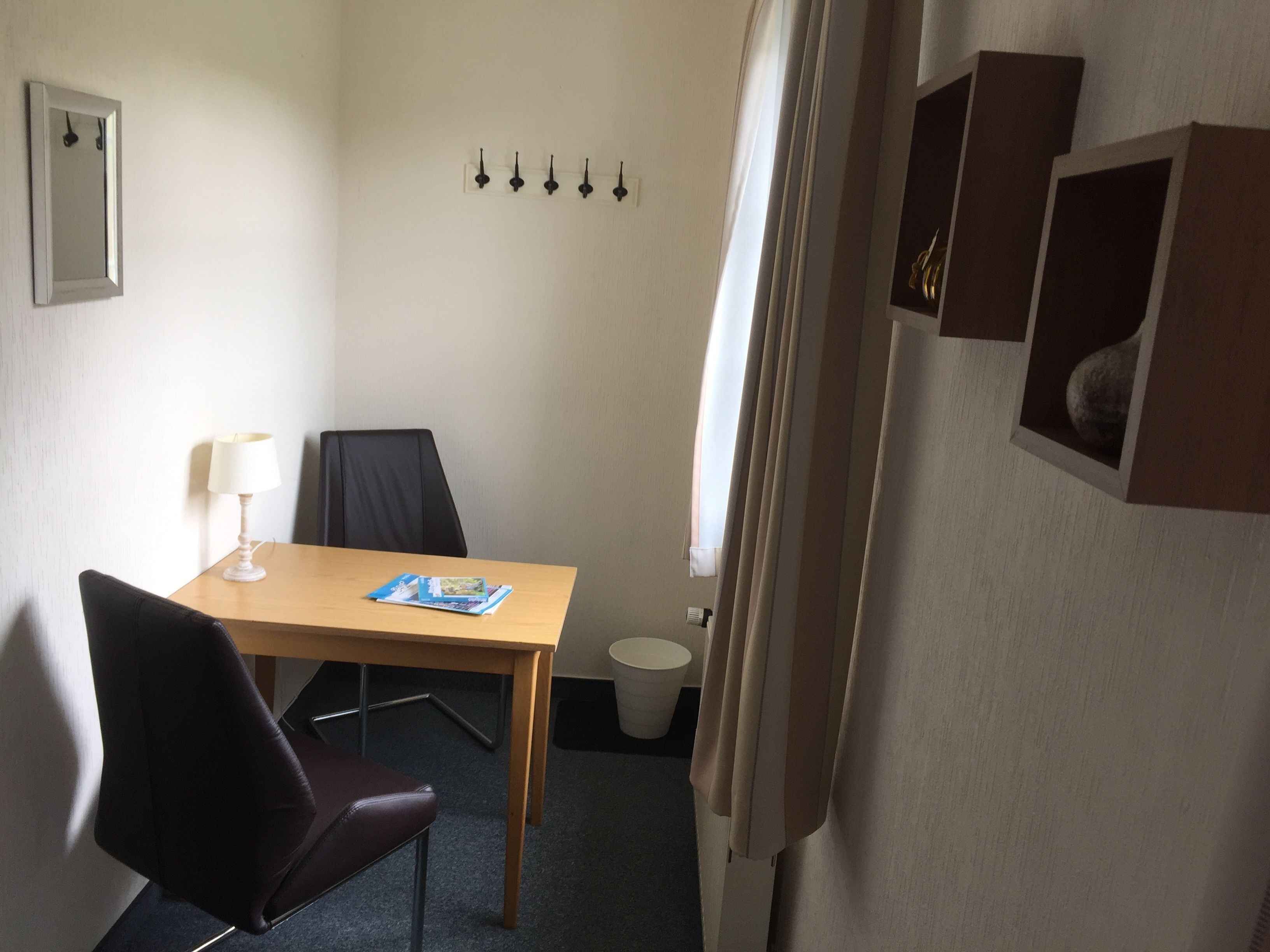 Una habitación pequeña y privada con una mesa de madera, dos sillas, una lámpara blanca, un ventanal con cortinas, un estante en la pared con libros y un inodoro blanco.