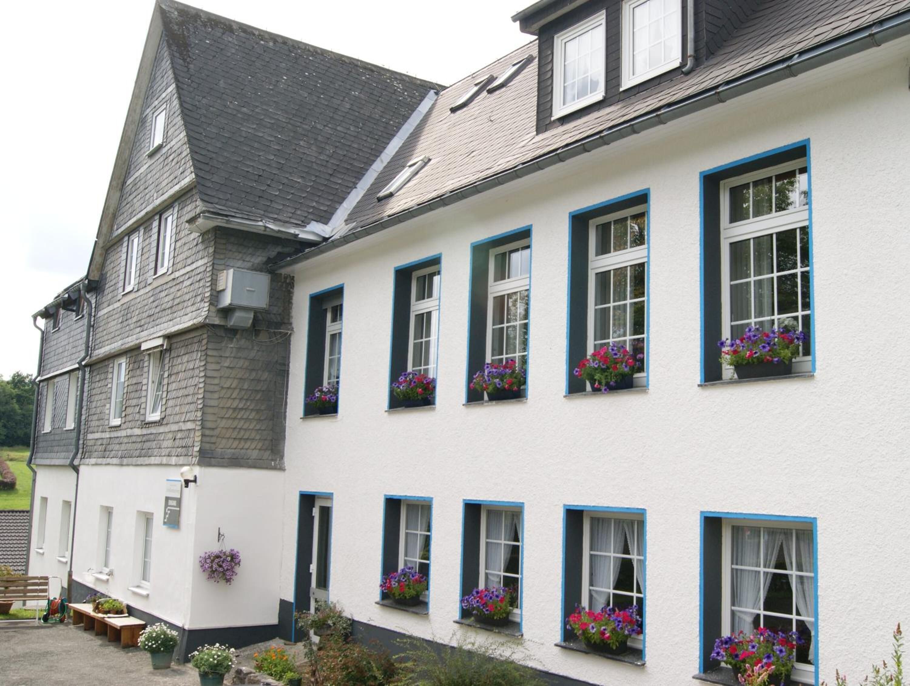 Een gebouw met twee verdiepingen, witte muren en blauwe raamkozijnen, voorzien van meerdere bloembakken met kleurrijke bloemen.