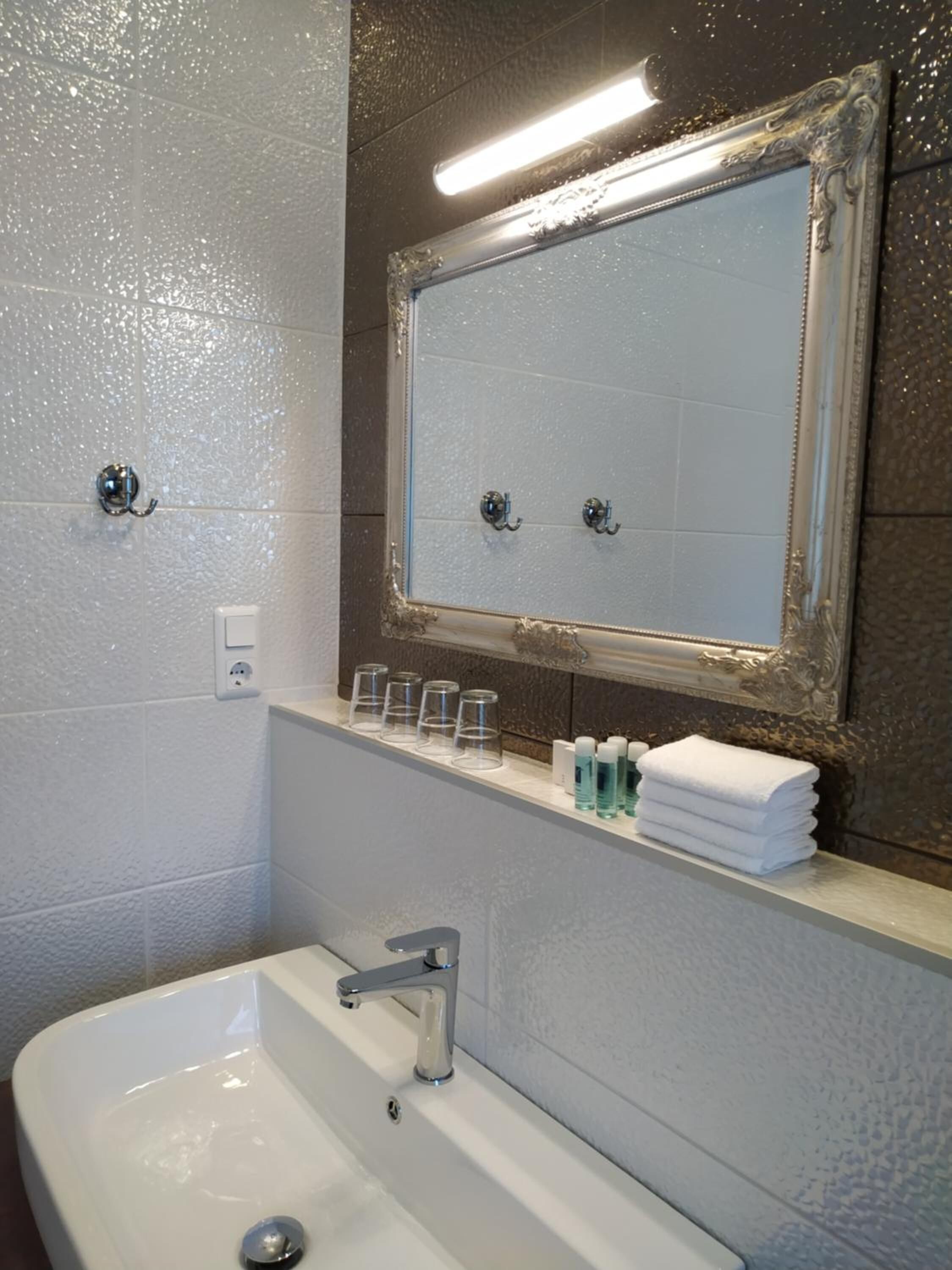 Une salle de bain avec un lavabo blanc, un miroir doté d’un cadre décoratif et une étagère contenant des articles de toilette.