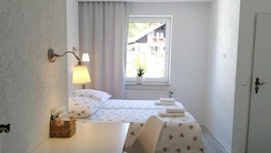 Habitación pequeña con cama, una mesa y dos sillas. Una ventana ofrece luz natural y vistas a edificios.