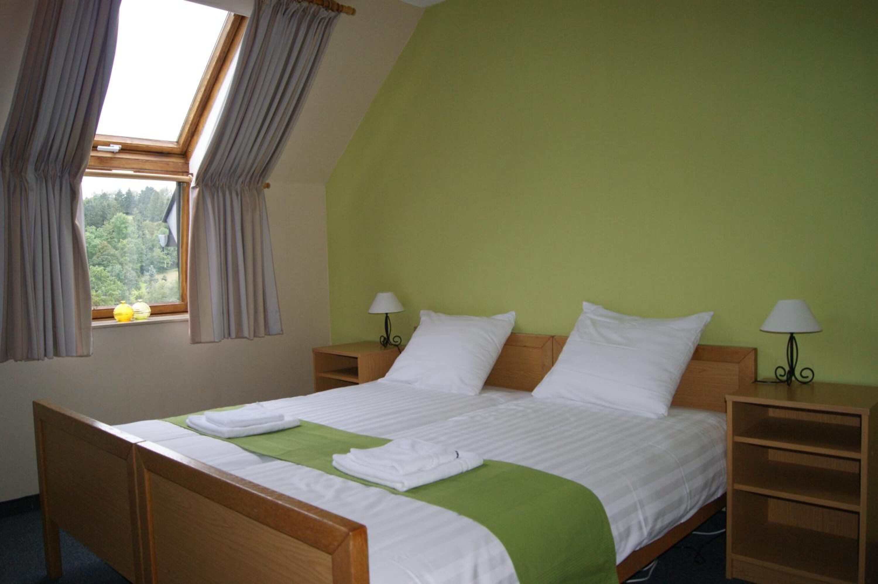 Una cama bien tendida con sábanas blancas y un plaid verde, dos lámparas de noche, una mesita de madera y una ventana con cortinas.
