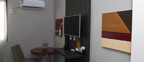 Una habitación pequeña con escritorio, silla, aire acondicionado, televisor de pantalla plana y una obra de arte en la pared.