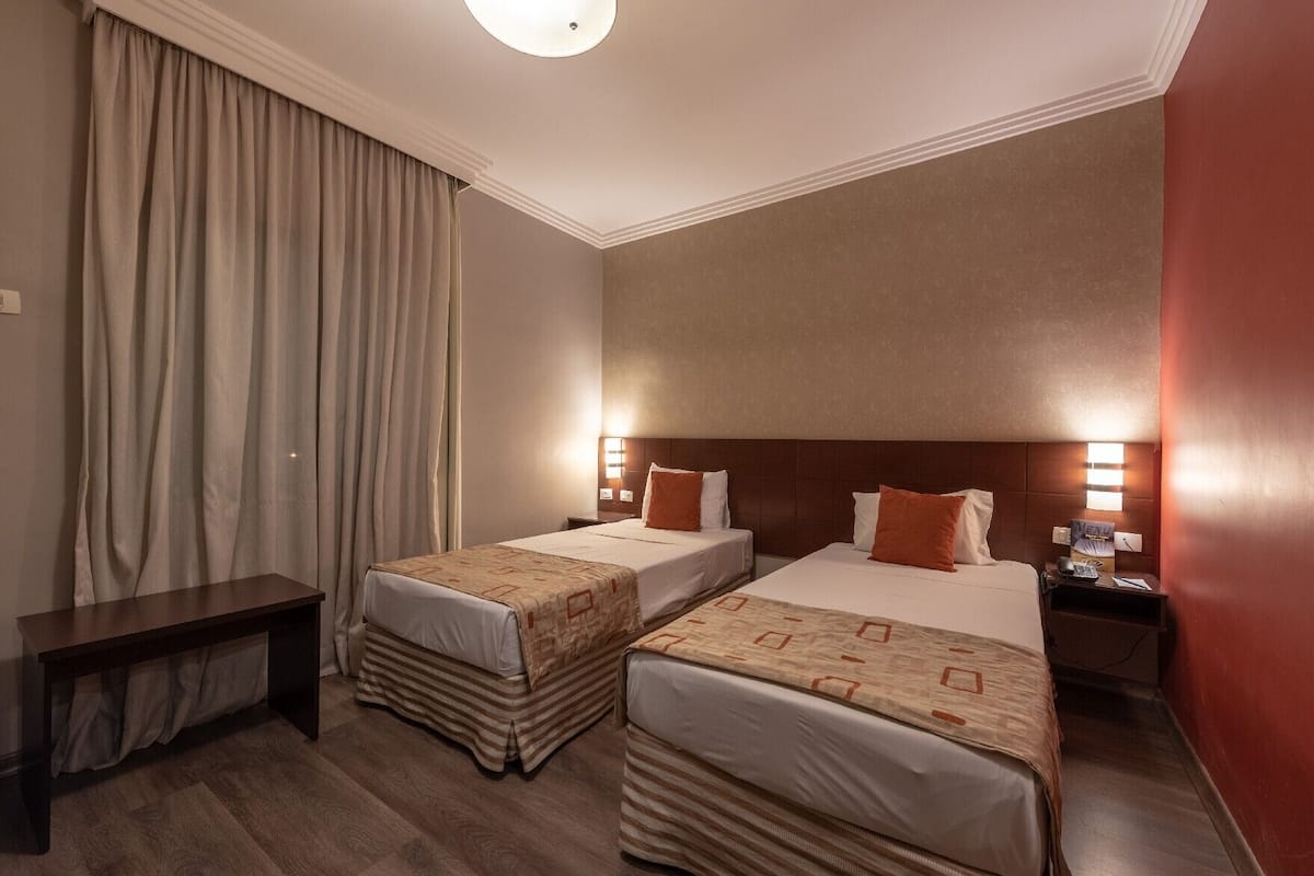 Une chambre d’hôtel avec deux lits, une table de chevet, un bureau et une grande fenêtre avec des rideaux.